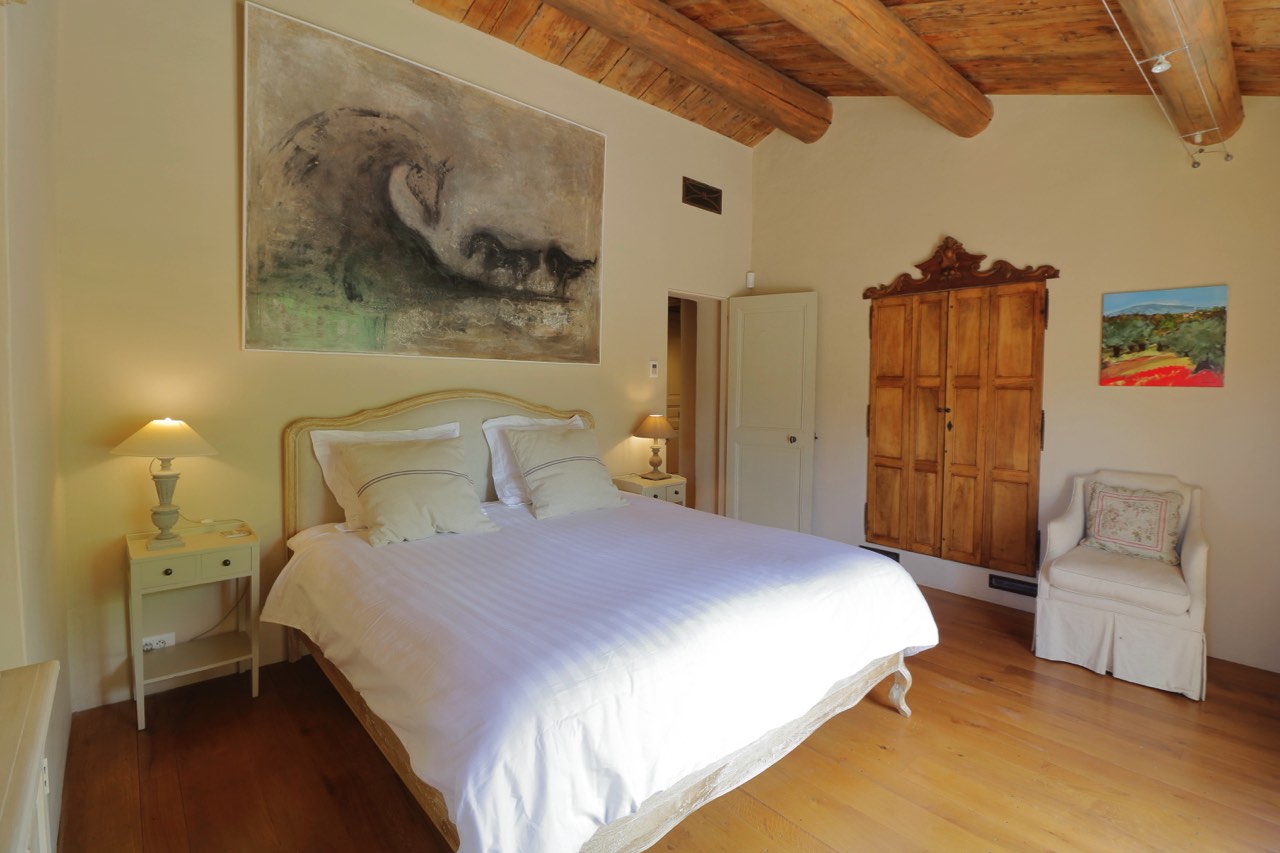 A cozy bedroom featuring a large bed with white bedding, rustic wooden furniture, and a large abstract painting above the bed. Exposed wooden beams add a traditional touch to the room.